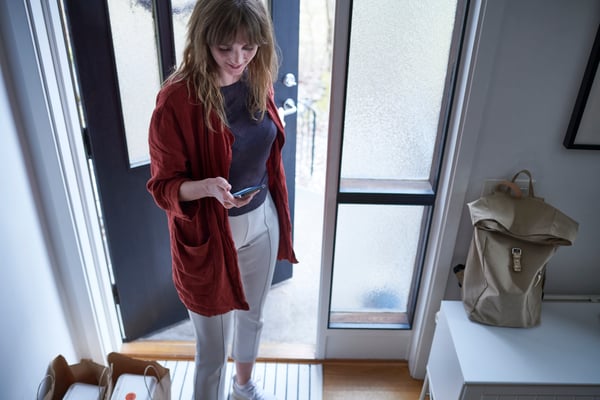 Une femme se tient devant sa porte d’entrée et regarde son appareil mobile.