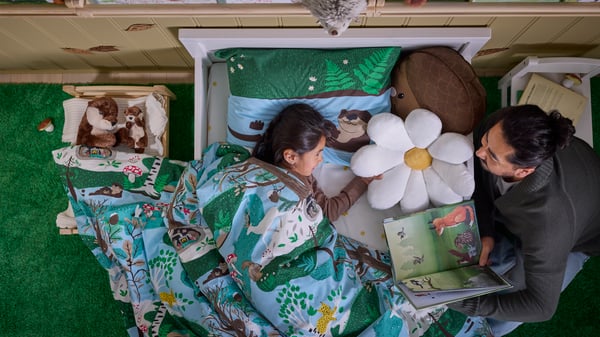 A child is lying in a white bed with SKOGSDUVA bed linen. In the bed is also a white SKOGSDUVA flower-shaped cushion.