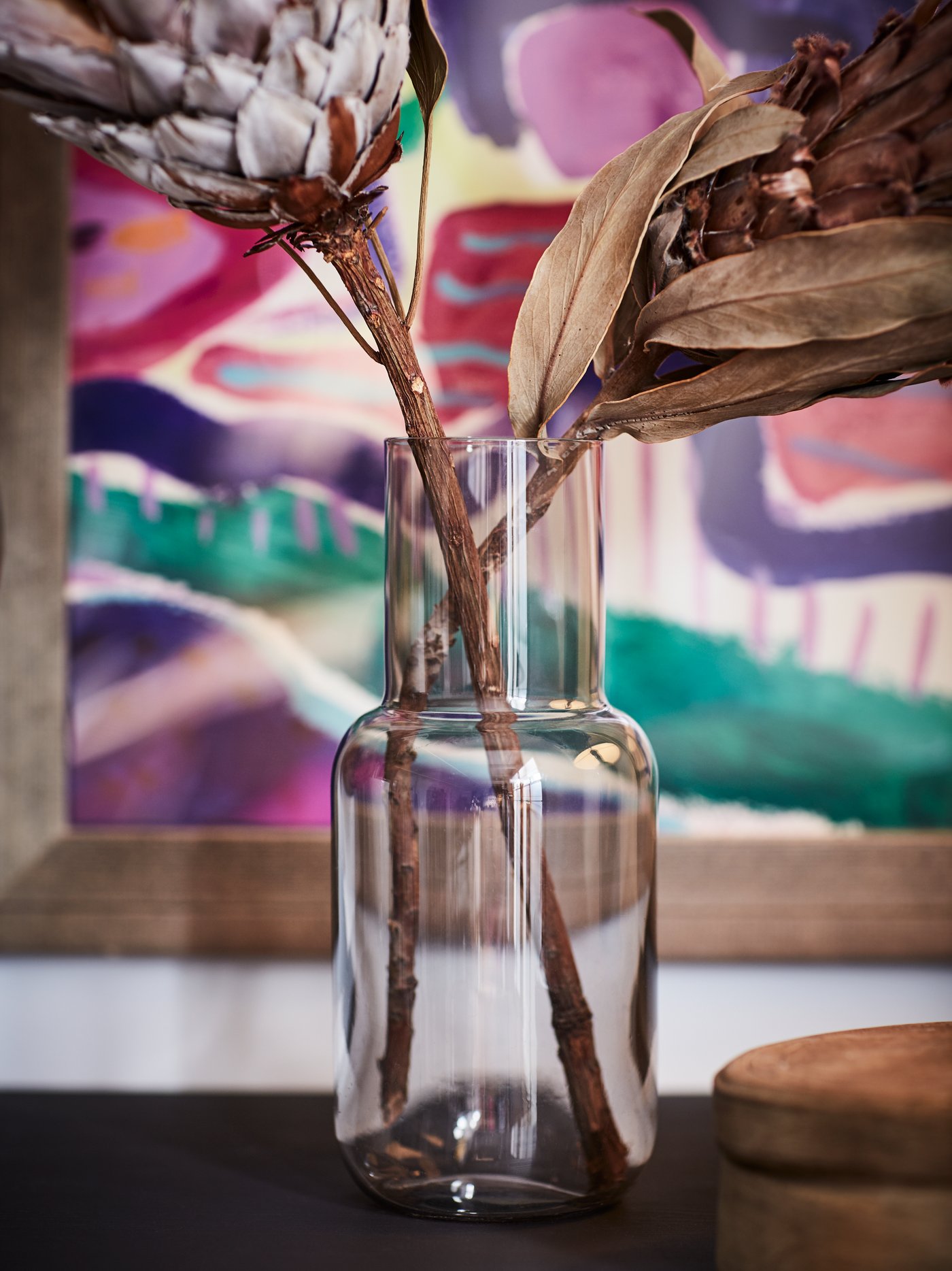 صورة مقربة لزهرتين مجففتين بسيقان سميكة في مزهرية زجاجية ولوحة ملونة في الخلفية.