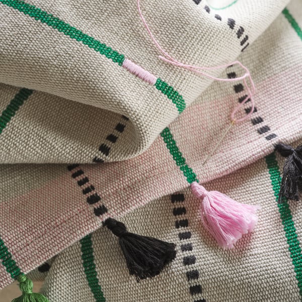 A close-up of the black and pink tassels of a handmade MÄVINN table-runner with stripes. On top is a needle with pink thread.