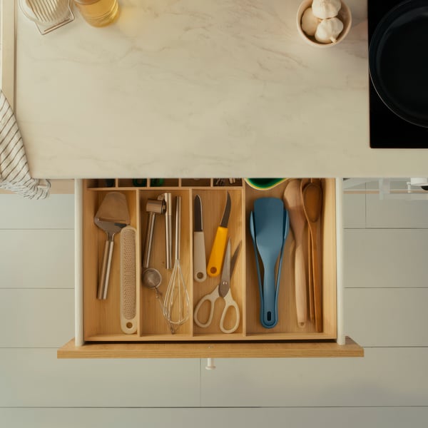 Un tiroir de cuisine bien rangé contenant divers ustensiles : spatules, fouet, ciseaux, épluche-légumes et cuillères en bois, séparés dans un organiseur en bois.
