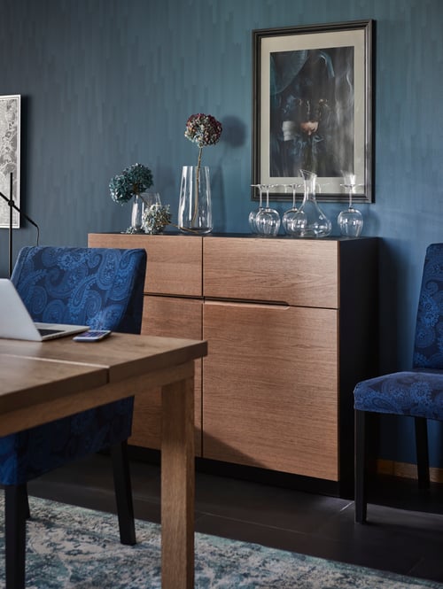 Ein braunes KLACKENÄS Sideboard an einer blauen Wand mit Glasvasen darauf. In ihnen ein paar Blumen. Daneben steht ein blauer Stuhl.