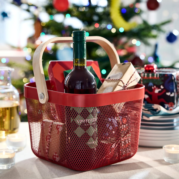 A red RISATORP basket filled with wrapped gifts and a bottle of VINTERSAGA mulled fruit drink and a gingerbread house.