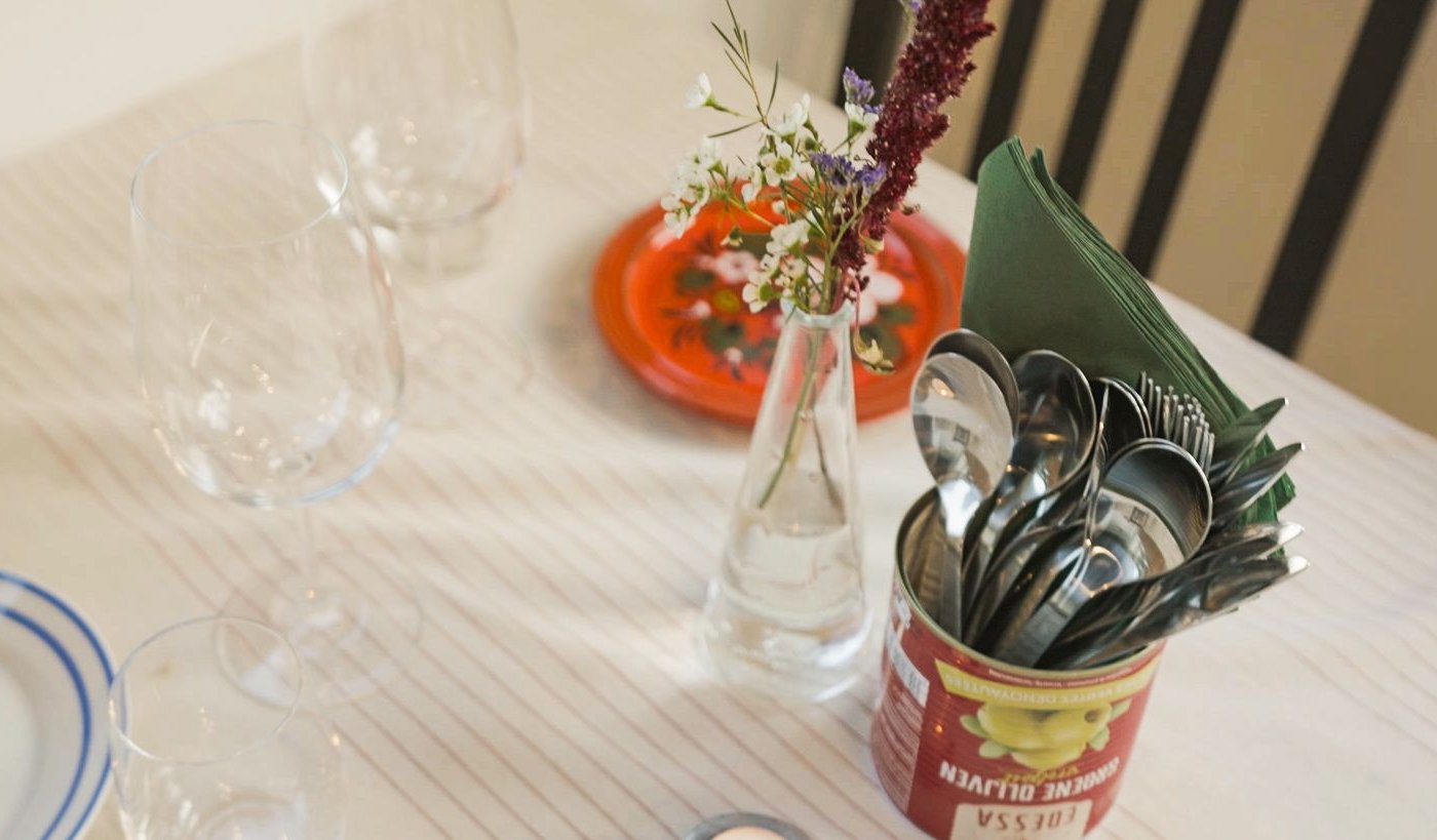 Tafel met glazen, wit bord met blauwe rand, bloemenvaas en bestek in rood blik. Sfeervol en creatief gedekt in gezellig restaurantinterieur.
