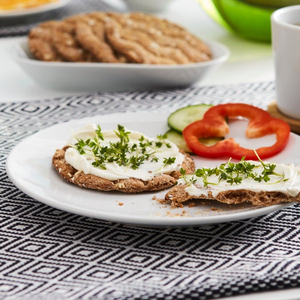 خبز مقرمش متعدّد الحبوب KNÄCKEBRÖD FLERKORN مع جبن كريمي وأعشاب على صحن أبيض فوق مفرش أطباق منقوش.