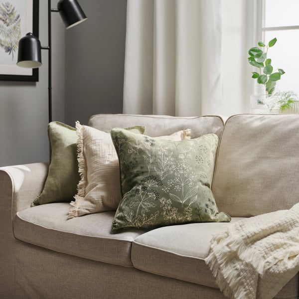 Un canapé IKEA confortable, agrémenté de coussins décoratifs verts et crème, d’un plaid tricoté douillet, de lampes murales noires et d’une plante verte, s’intègre dans un intérieur lumineux et apaisant.