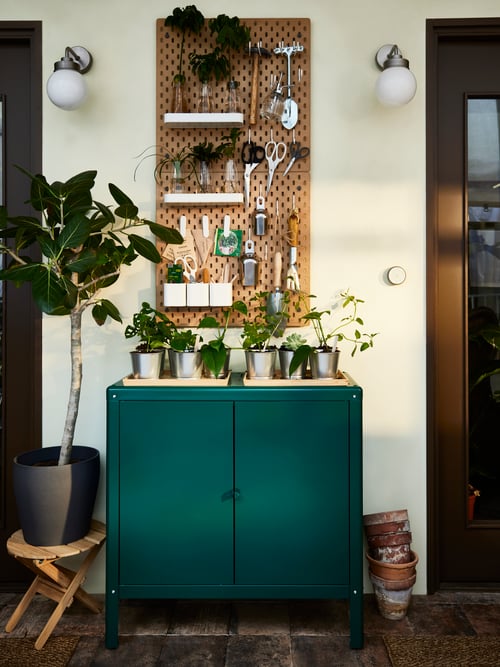 SKÅDIS pegboards with gardening items, a green steel cabinet with seedlings on top, two FRIHULT wall lamps and a big plant.