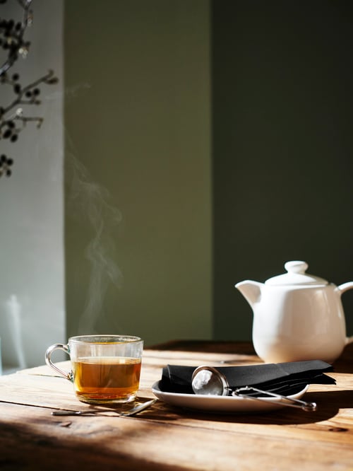 Dining area with wooden table, clear glass mug with tea, tea infuser, white teapot, side plate, coffee spoon, paper napkin.