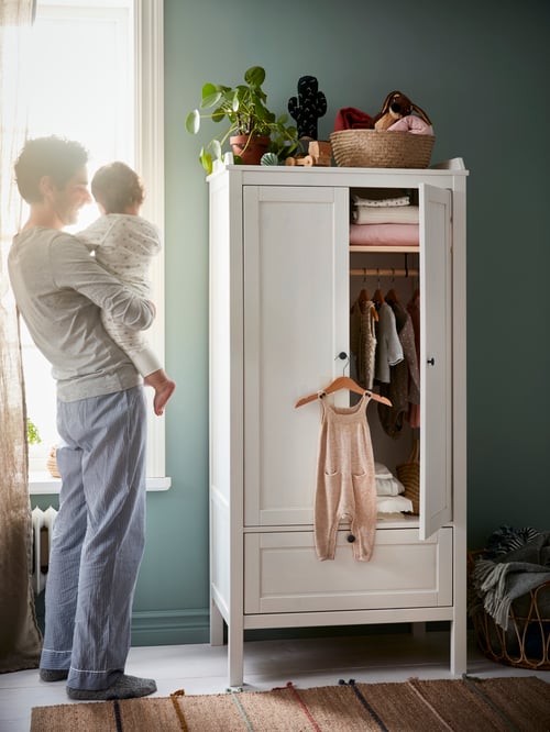 A man holding a baby looks out a window by a white SUNDVIK wardrobe. An open door above a drawer shows baby clothes inside.