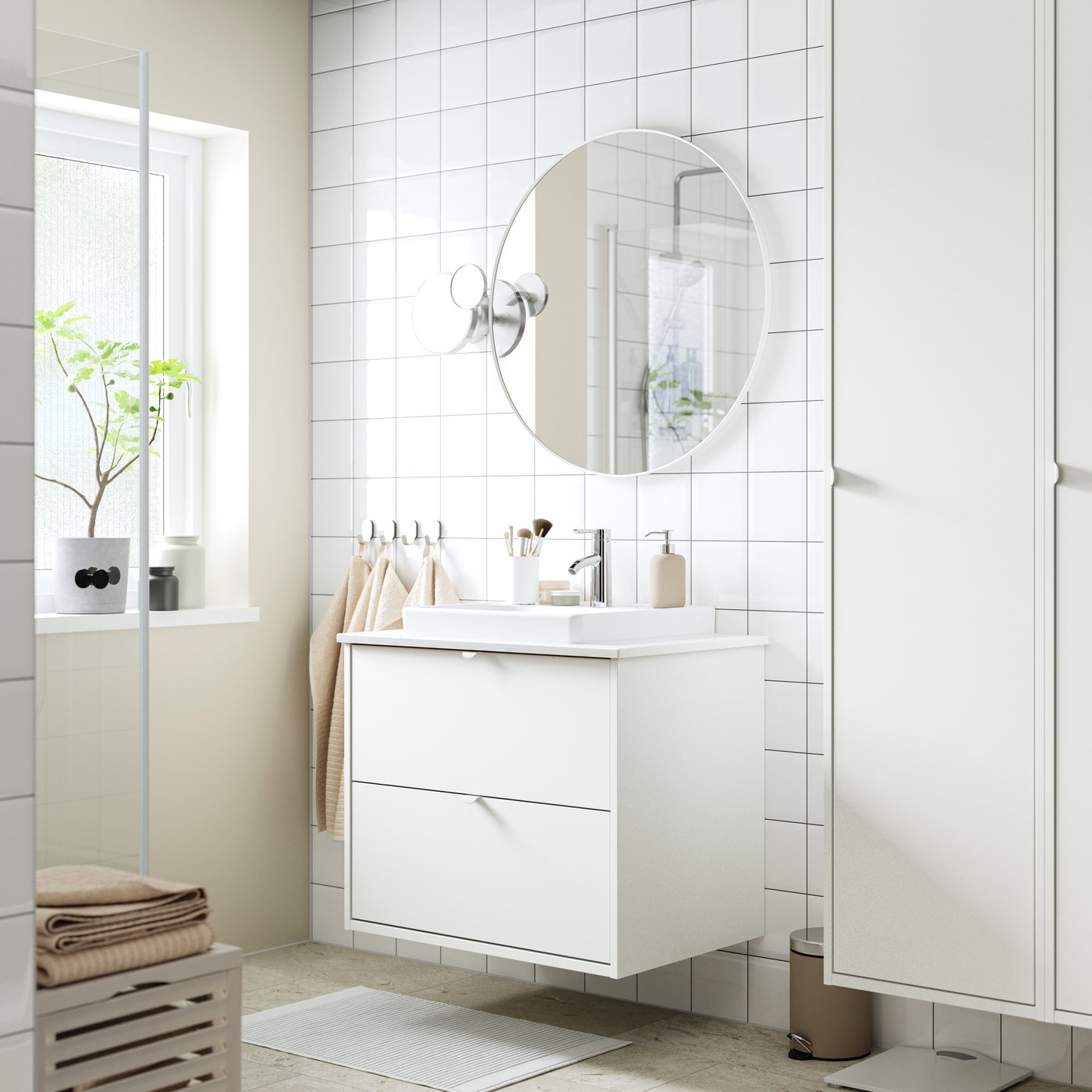 Osvetljeno, potpuno belo kupatilo s belim HAVBÄCK postoljem umivaonika s fiokama pored odgovarajućeg visokog elementa i zaobljenog ogledala.