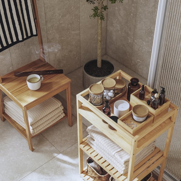 A sunlit corner of a bathroom with RÅGRUND trolley and a towel rack chair in natural bamboo filled with spa essentials.