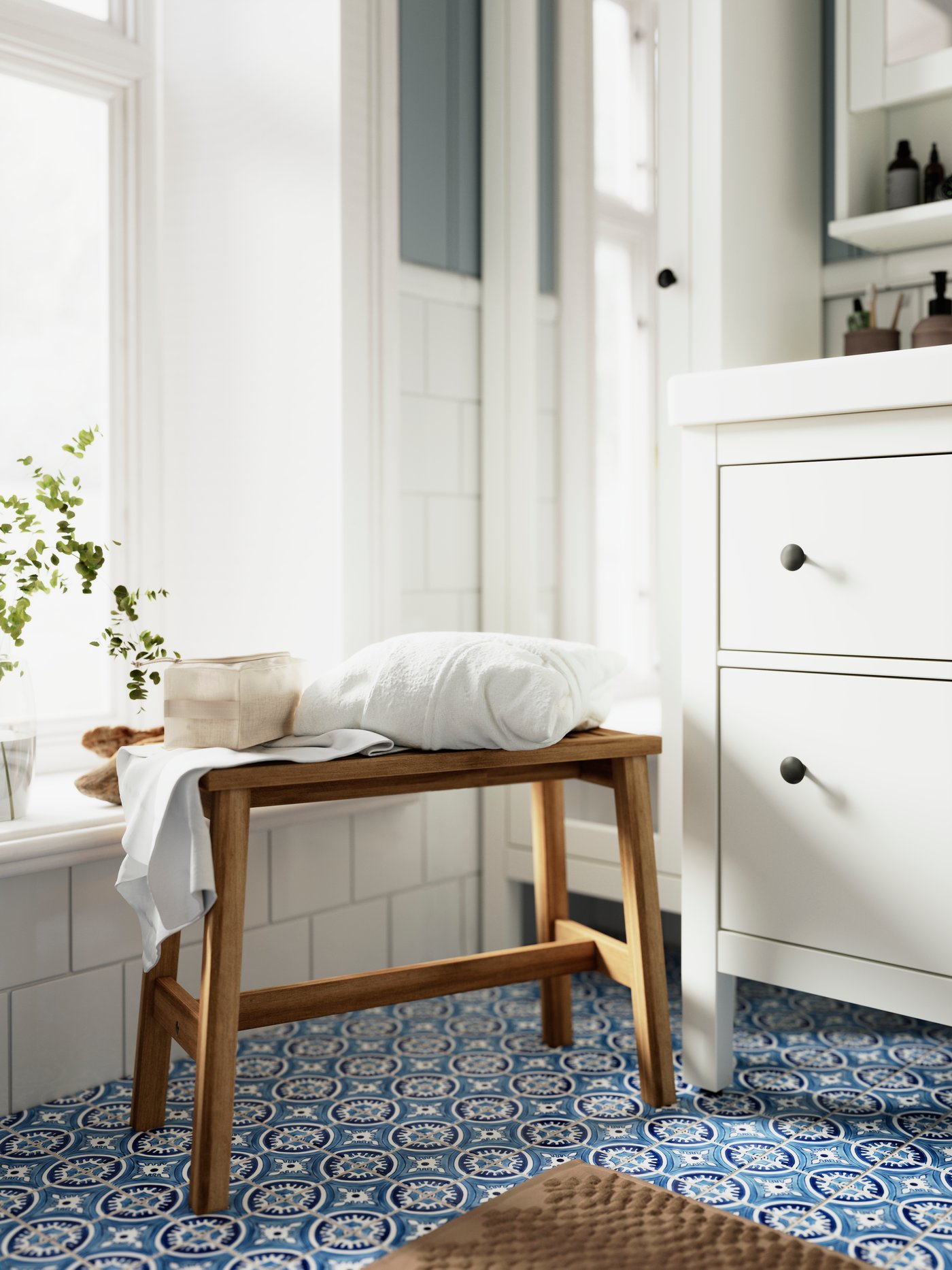 Ett ljust, vitkaklat badrum med blått mosaikgolv har vita HEMNES badrumsmöbler och en STOREDAMM-bänk i trä.