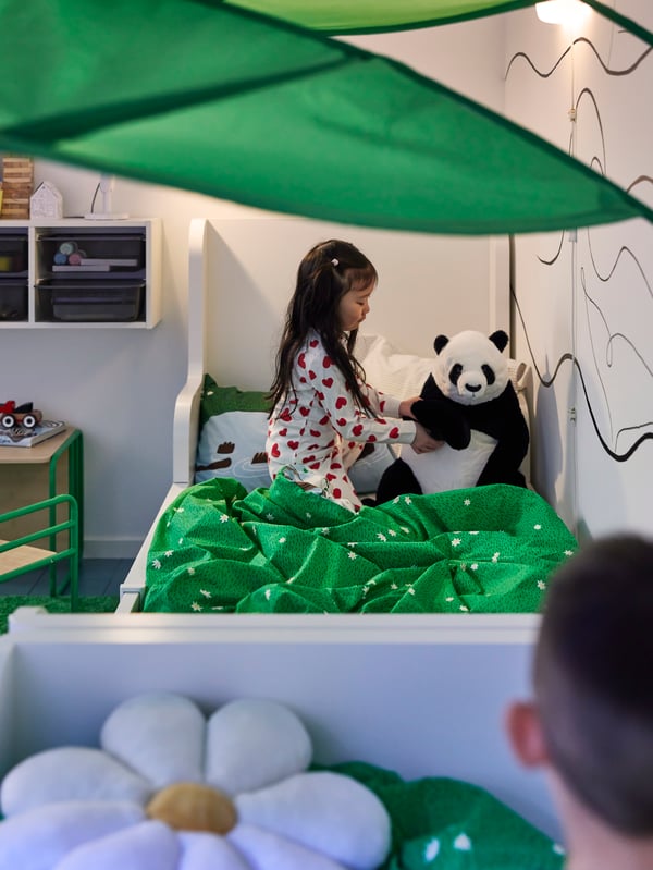 Liebevoll eingerichtetes IKEA Kinderzimmer mit einem weißen Bett und einer grünen Decke. Ein Mädchen im Pyjama spielt mit einem großen Panda-Stofftier. Das grüne Blätterdach sorgt für ein verspieltes, naturnahes Ambiente im Kinderzimmer.

