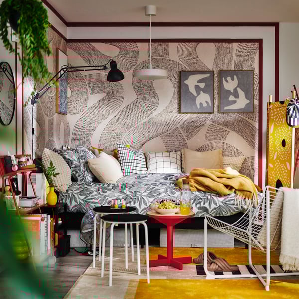 Two MARIUS stools and a red side table by a SLATTUM upholstered bed with BROKDRACENA patterned bed linen and cushions on top.