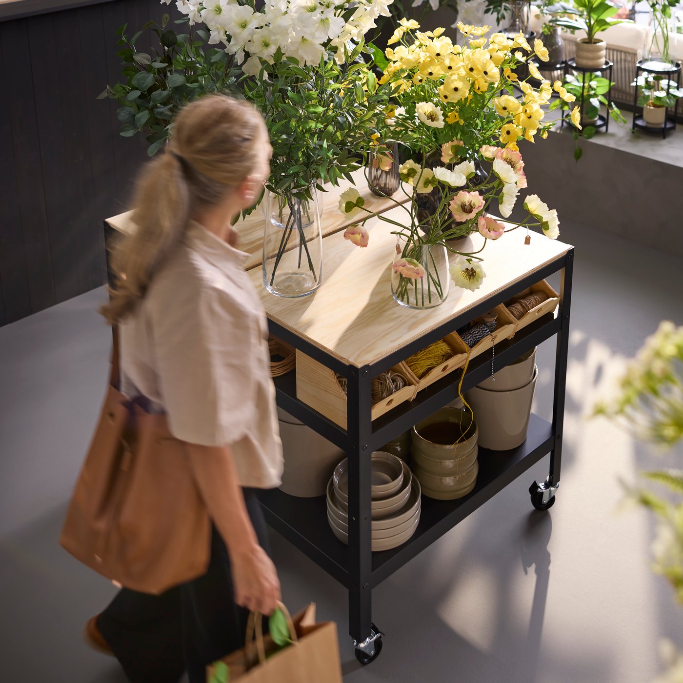 A person in a flower shop scene walking by a black BROR trolley made from pine plywood, holding plants and flowers.