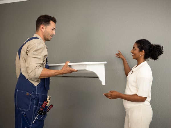 A man in work overalls holds a white shelf while a woman points to a grey wall, seemingly discussing its placement.