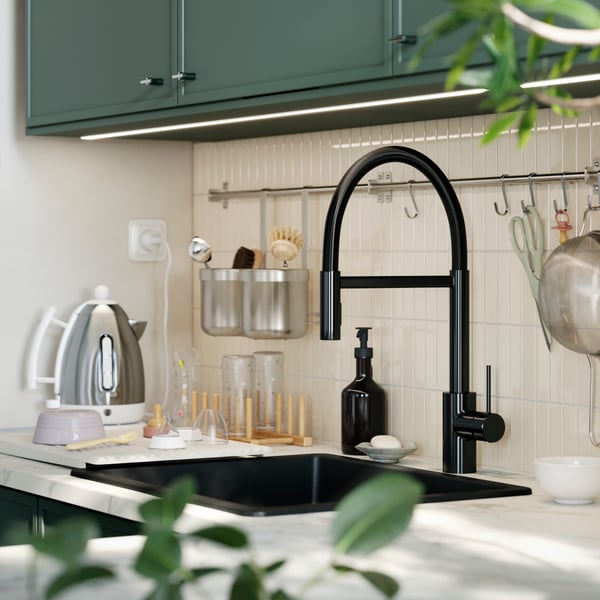 A SALLSJÖN kitchen mixer tap with a gooseneck design is mounted in a kitchen with a tiled wall and grey-green kitchen doors.