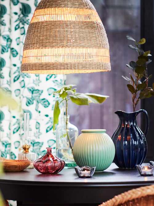 A handwoven, seagrass TORARED pendant lampshade hangs above a round table that has various vases and tealights on top.