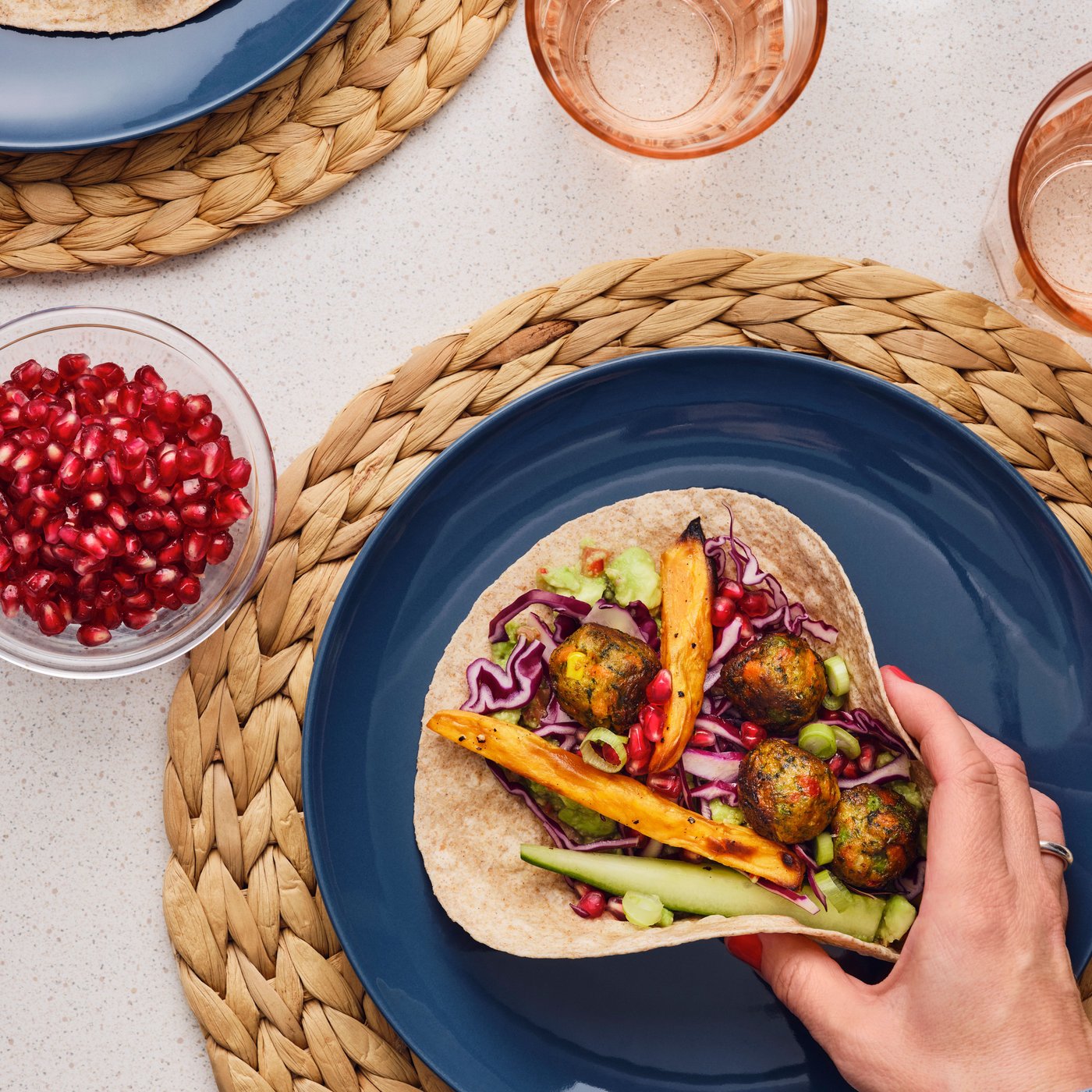 Une personne prend une tortilla remplie de boulettes de légumes et de légumes colorés, placée sur une assiette bleue.