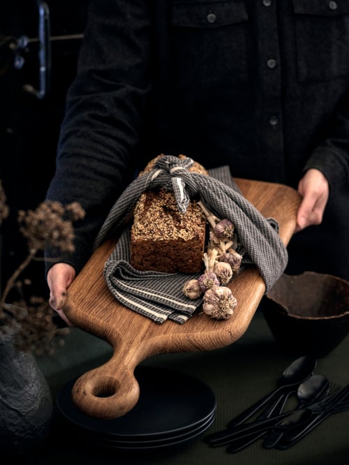 A person presents an oak ARTISTISK chopping board with garlic and homemade bread wrapped in a grey MARIATHERES tea towel.