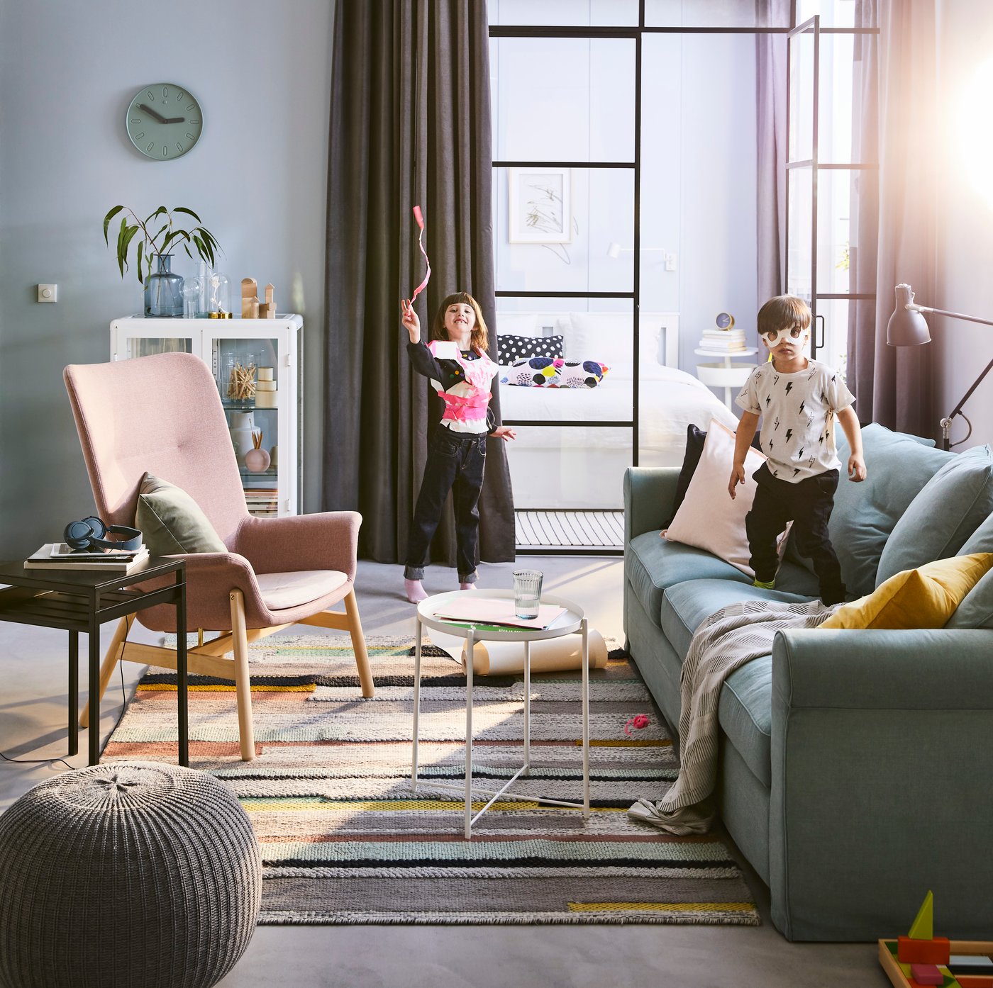 2 Kinder spielen in einem Raum und haben spaß. Ein Kind springt auf einem grauen Sofa. Vor dem Sofa stehen 2 kleine Tische und ein rosaner Stuhl.