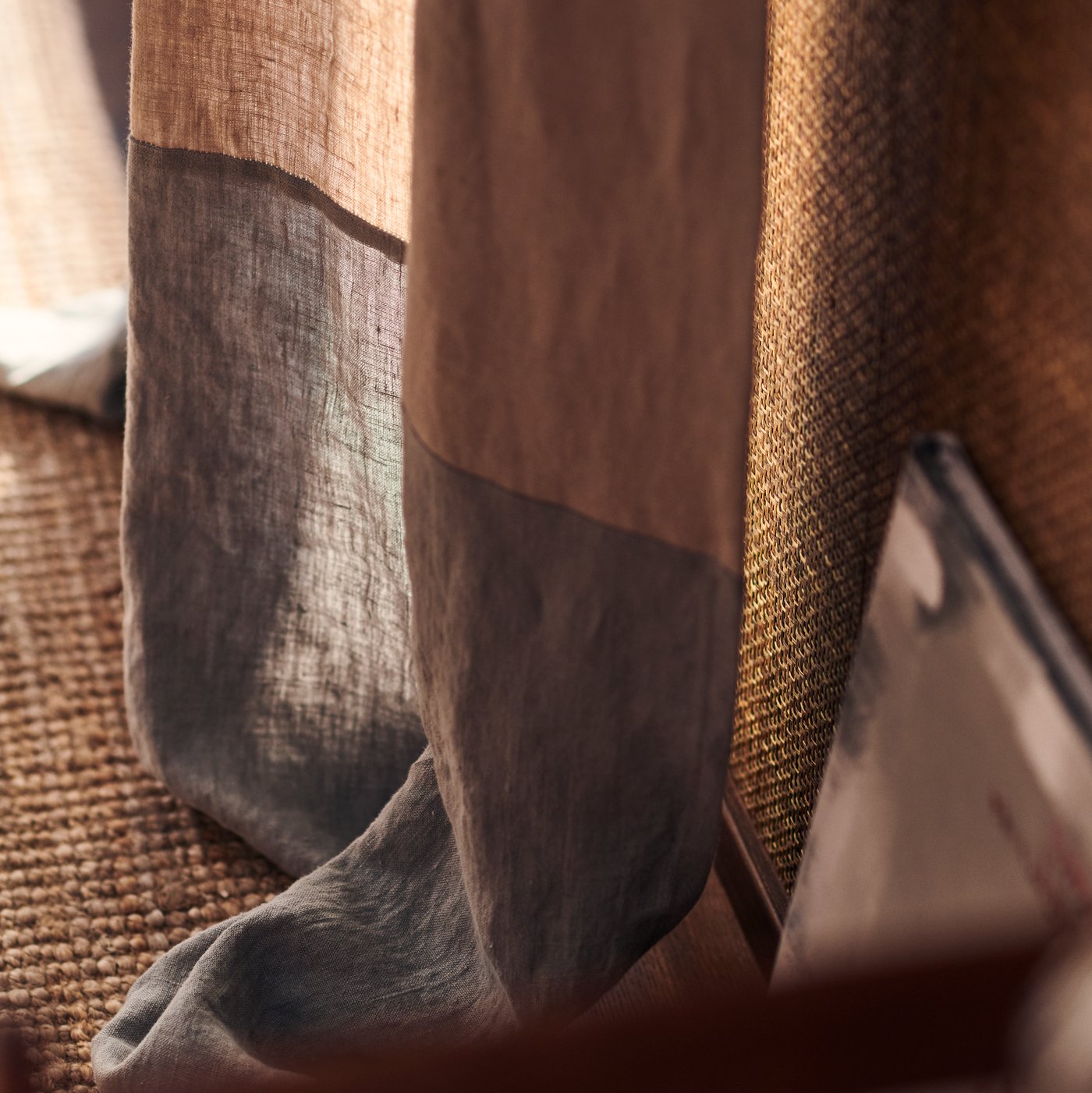 الجزء السفلي من ستارة ملونة باللون البني والرمادي، متدلية فوق سجادة LOHALS مصنوعة من الأعشاب