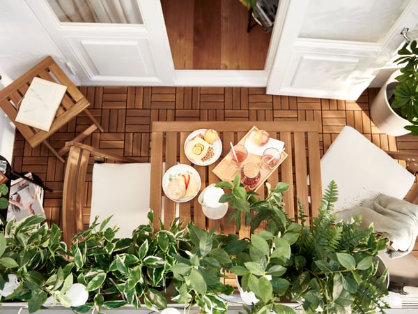 A balcony characterised by slatted acacia – NÄMMARÖ furniture and RUNNEN floor decking – and leafy FEJKA artificial plants.