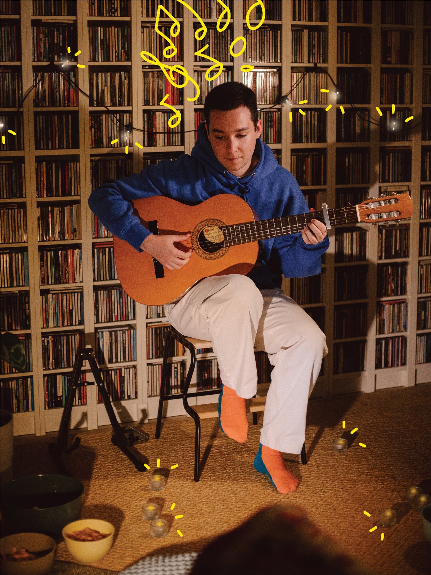 A man playing the guitar in front of a bookcase wall decorated with string lights.