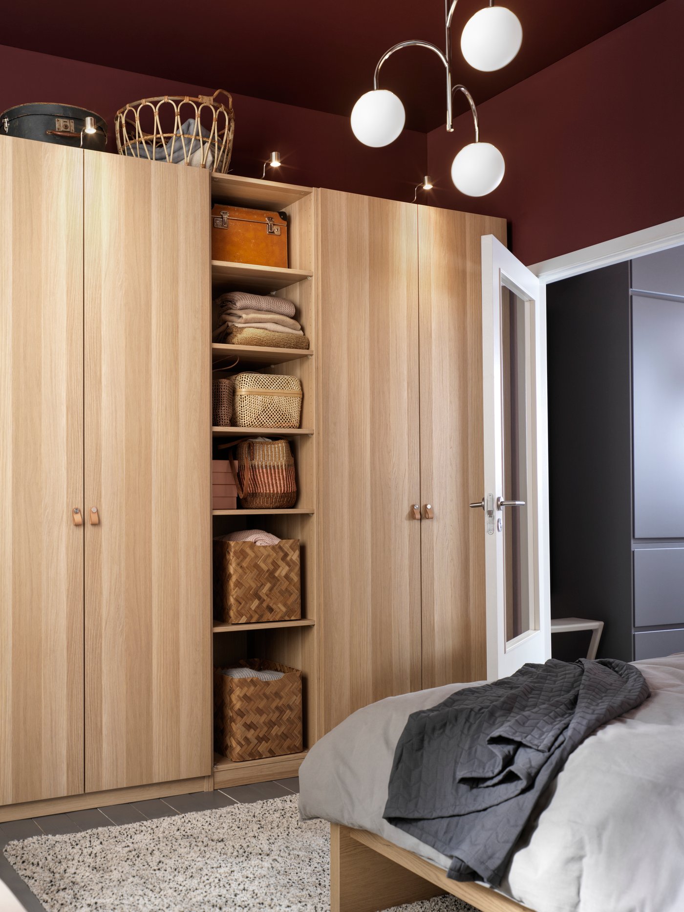 Dormitorio con varias puertas de madera en beige, una lámpara colgante de 3 brazos, una cesta redonda y una colcha gris.