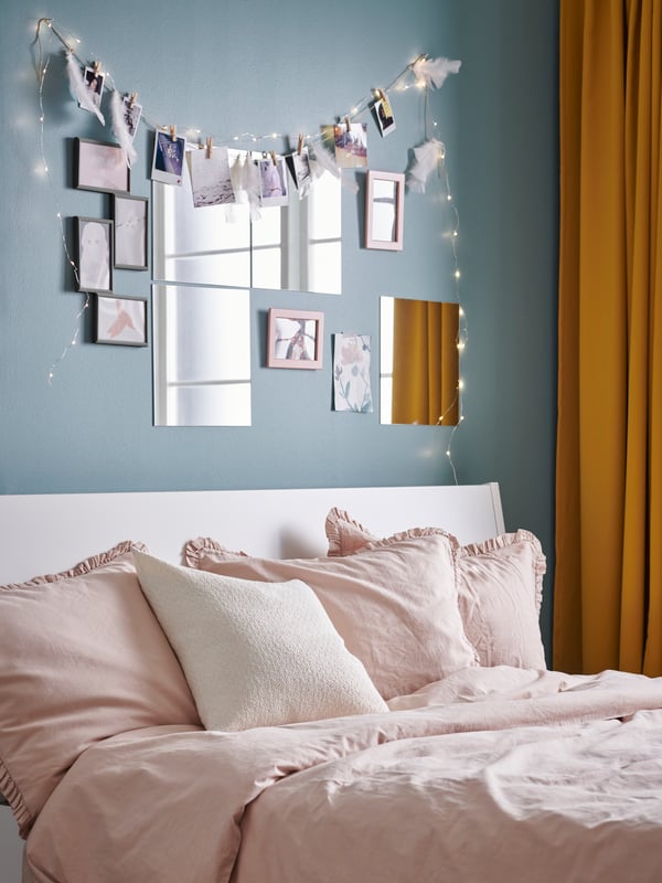 Un angolo di una stanza dalle pareti azzurre e con delle tende arancioni. Il letto ha delle lenzuola rosa e un cuscino bianco. Sulla parete azzurra degli specchi. 