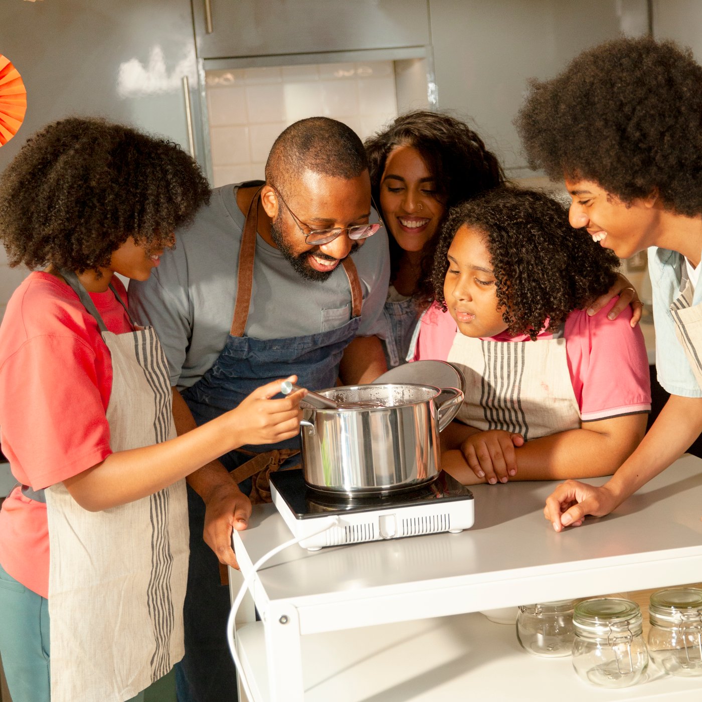 Una famiglia di cinque persone intente a mescolare il contenuto di una pentola IKEA 365+ su un piano cottura a induzione portatile TILLREDA.