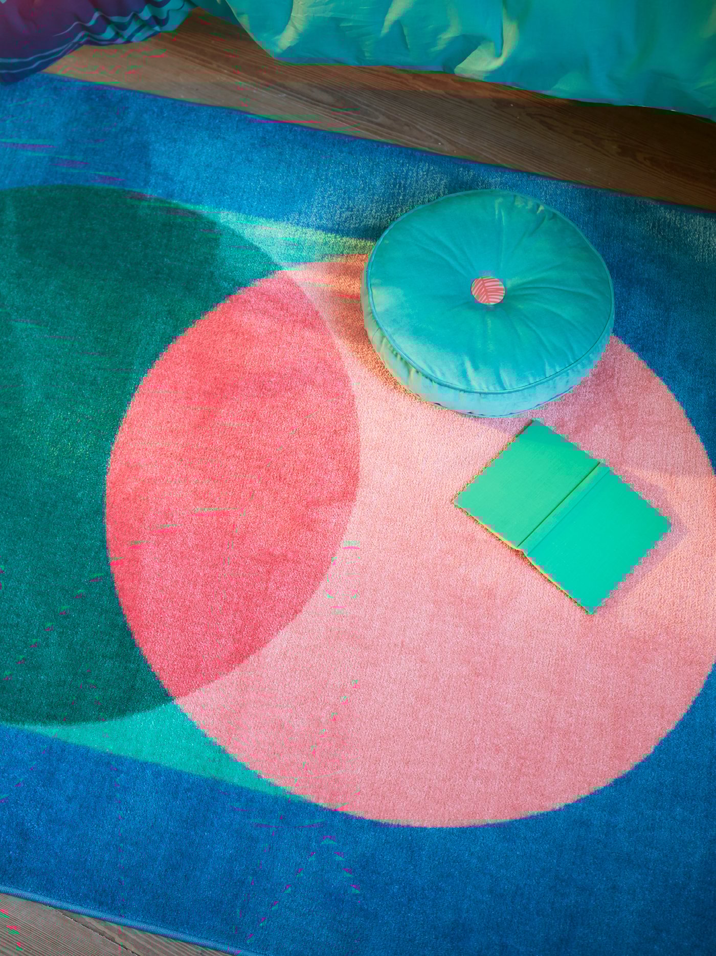 Un tapis GRACIÖS avec un grand motif circulaire en rose et vert sur fond bleu, avec un livre et un coussin posés dessus.