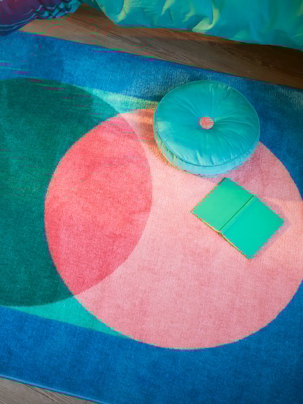 Alfombra GRACIÖS con un gran estampado circular en verde y rosa sobre un fondo azul, con un libro y un cojín encima.