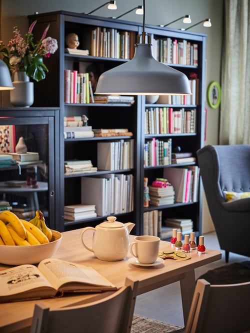 A dining table stands within a living room by a wall with two HEMNES black-brown bookcases that have cabinet lighting on top.