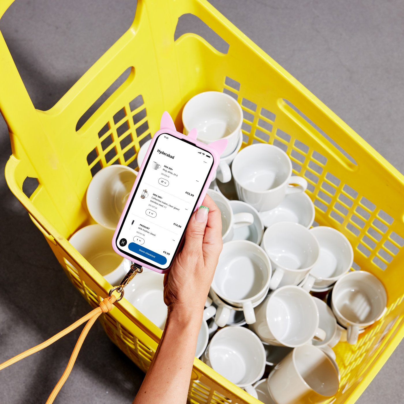 A hand holds a smartphone over a yellow shopping basket filled with white cups, displaying a shopping list page in the IKEA app.