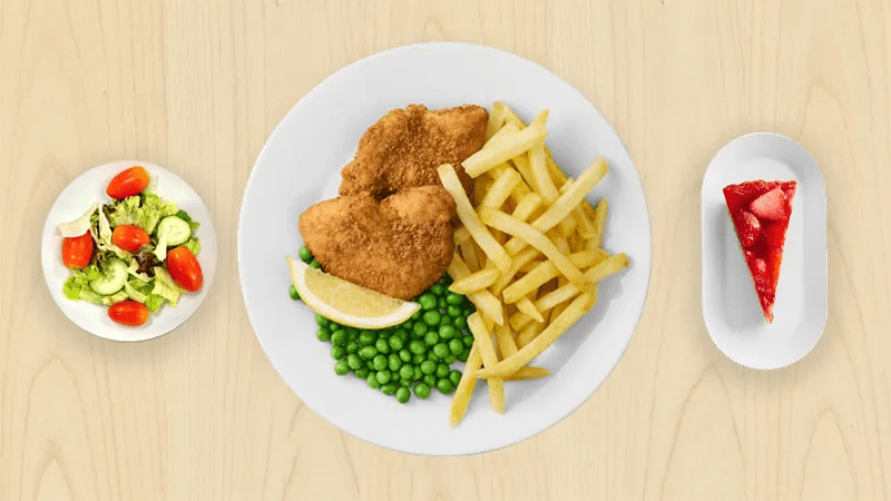 Puukuvioista taustaa vasten kolme lautasta, joissa kana schnitzel, pieni salaatti ja mansikkajuustokakkupala.