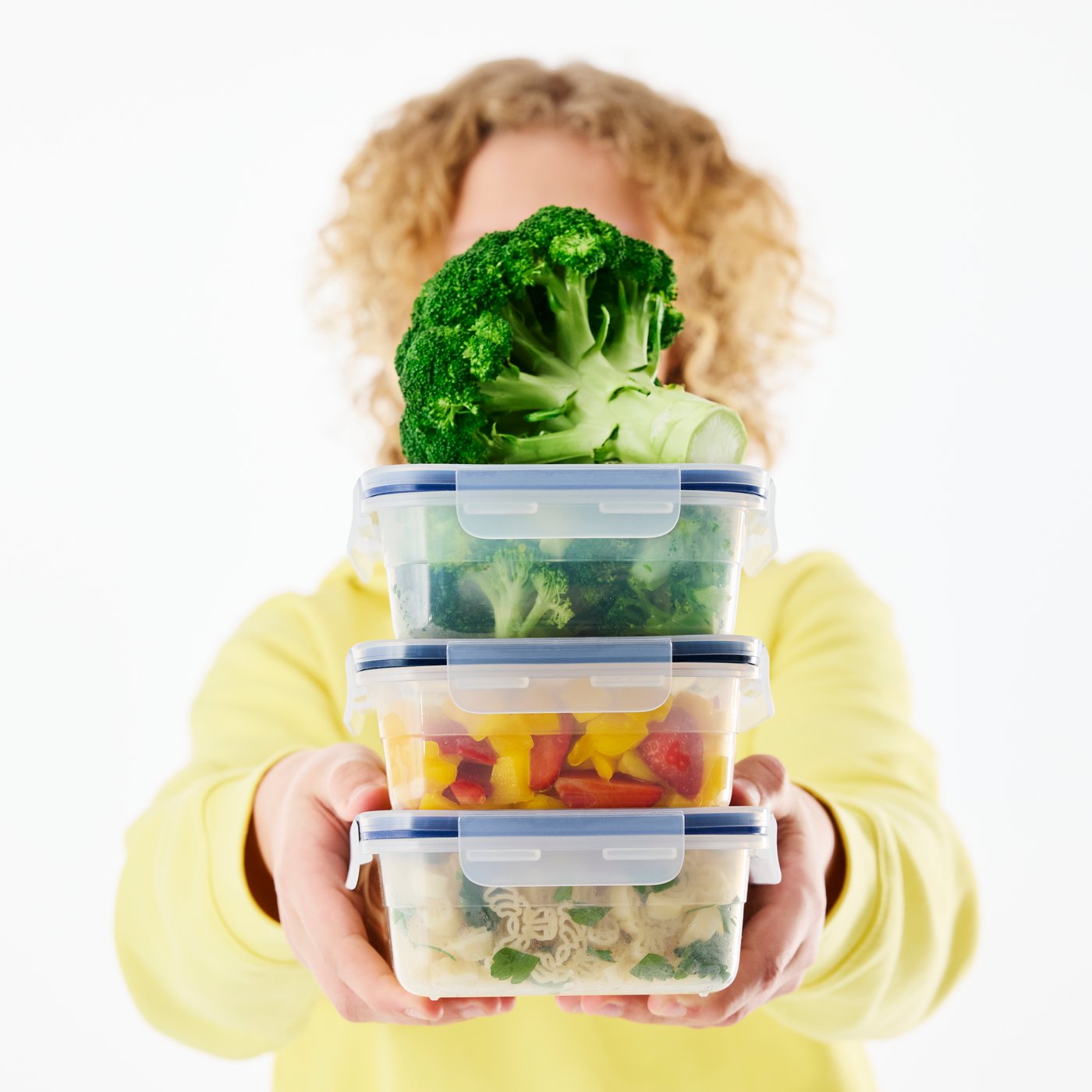 Una persona tiene in mano una pila di tre contenitori per alimenti in plastica IKEA 365+ pieni di cibo. Sopra c'è un broccolo crudo.