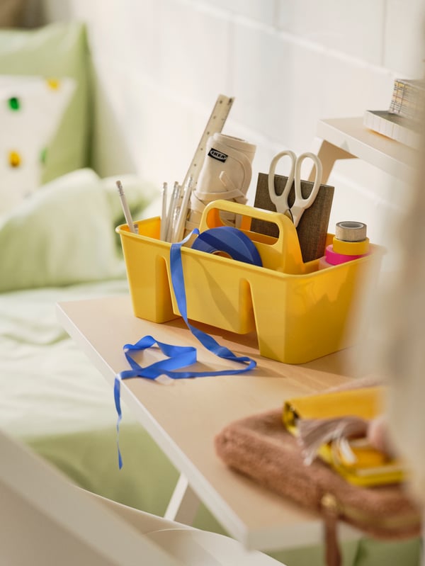 Close-up of small yellow portable storage with handles filled with stationary on white student desk in college dorm room.
