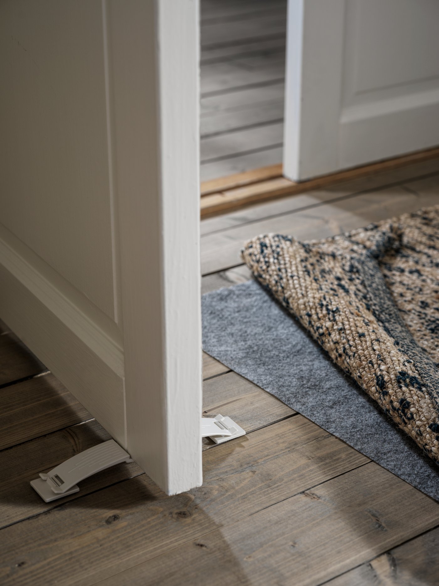 Detalle de un tope de puerta PATRULL sosteniendo una puerta blanca abierta. Junto a la puerta hay una alfombra MELHOLT adherida al piso de madera con un protector antideslizante STOPP FILT.
