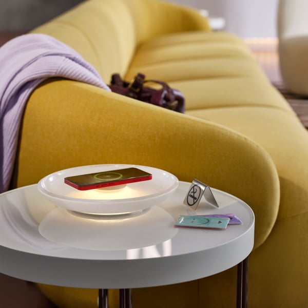 By a green-yellow sofa is a white table holding a donut-shaped VÄSTMÄRKE wireless charger with lighting by hotel key cards.