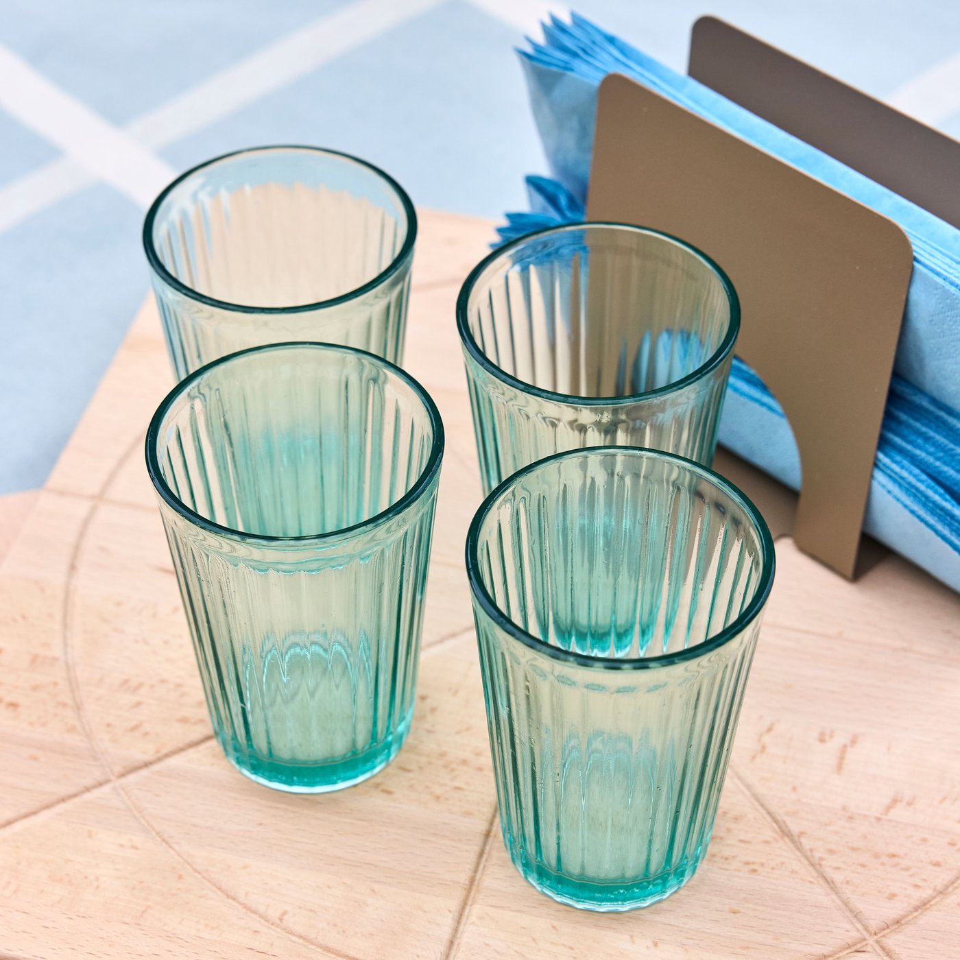 Vier geribbelde drinkglazen in lichtgroen glas op een houten tafel, naast een servethouder met blauwe servetten.
