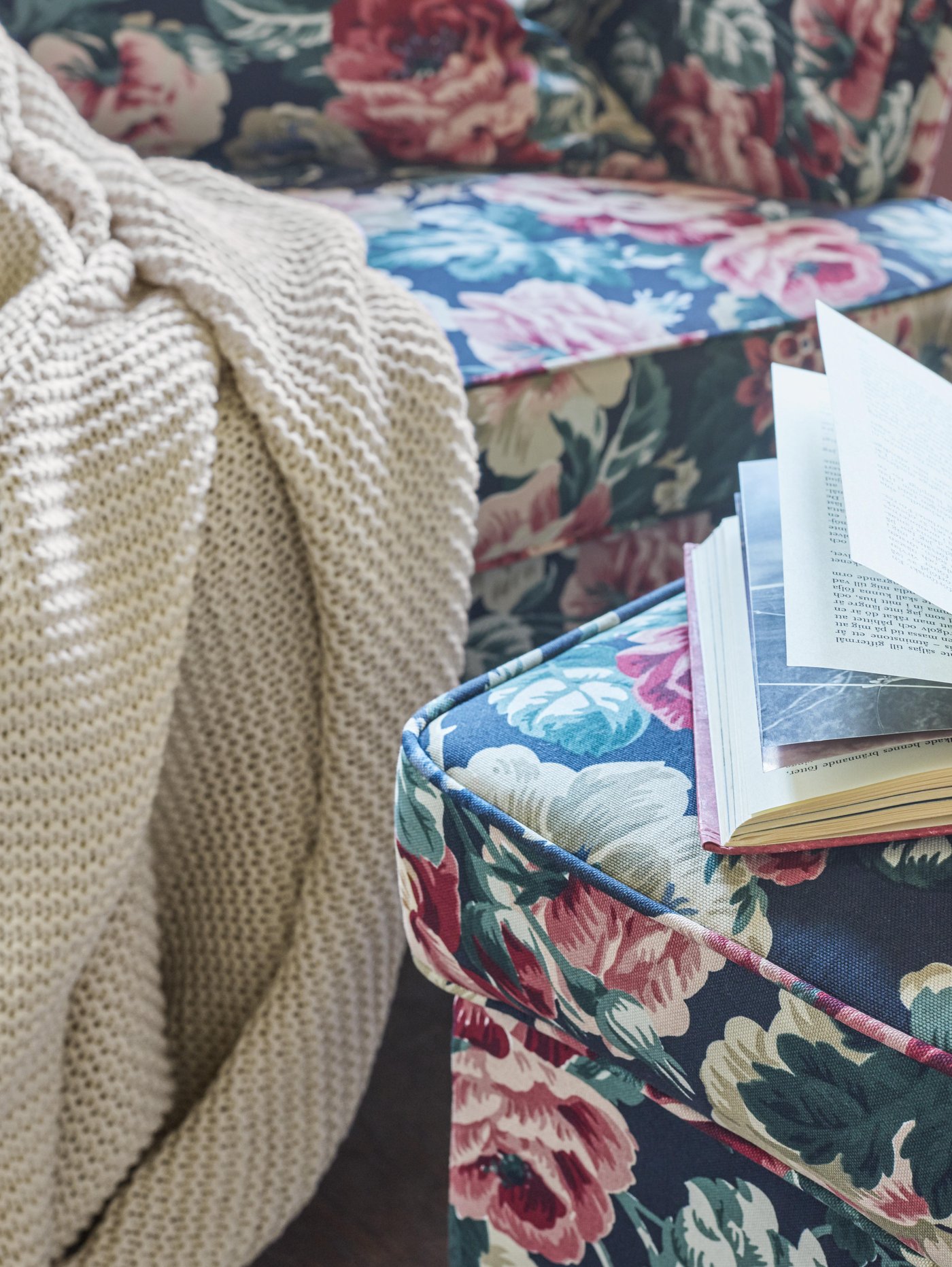 Light coloured INGABRITTA throw draped over the edge of a floral patterned sofa, near a footstool with an open book on it.