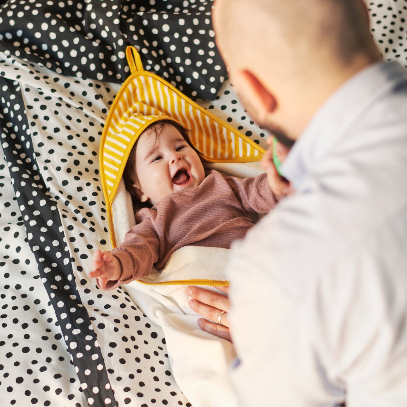 A baby is snuggled in a soft yellow GRÖNFINK towel with a hood, lying comfortably on a patterned bedspread.