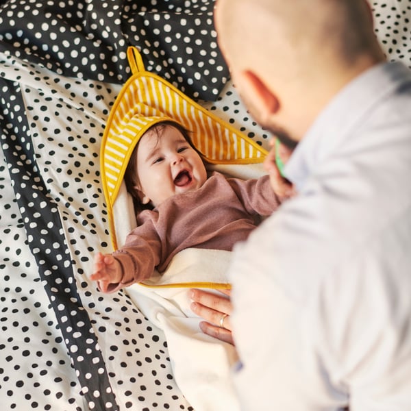 A baby is snuggled in a soft yellow GRÖNFINK towel with a hood, lying comfortably on a patterned bedspread.