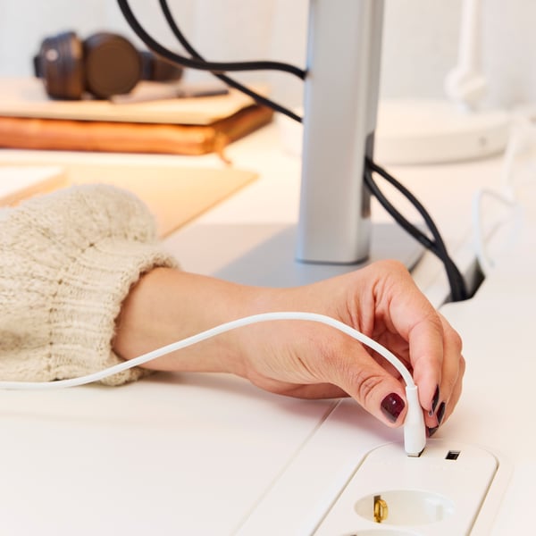 A person putting a USB cable in a white KOPPLA 4-way socket with 2 USB ports that is on top of a MITTZON sit/stand desk.