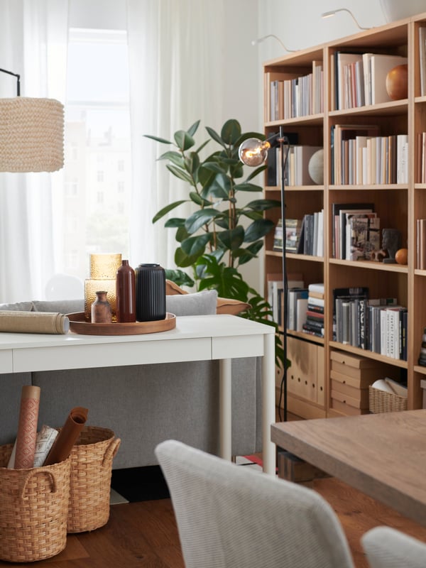 A bright living space with TONSTAD bookshelves, a potted plant placed by the window and a white desk with a tray with vases.
