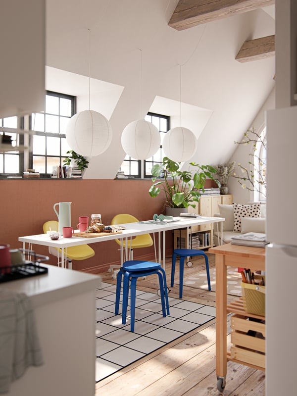 A sun-drenched dining area with two tables, blue stools, yellow chairs and three pendants made from natural materials.