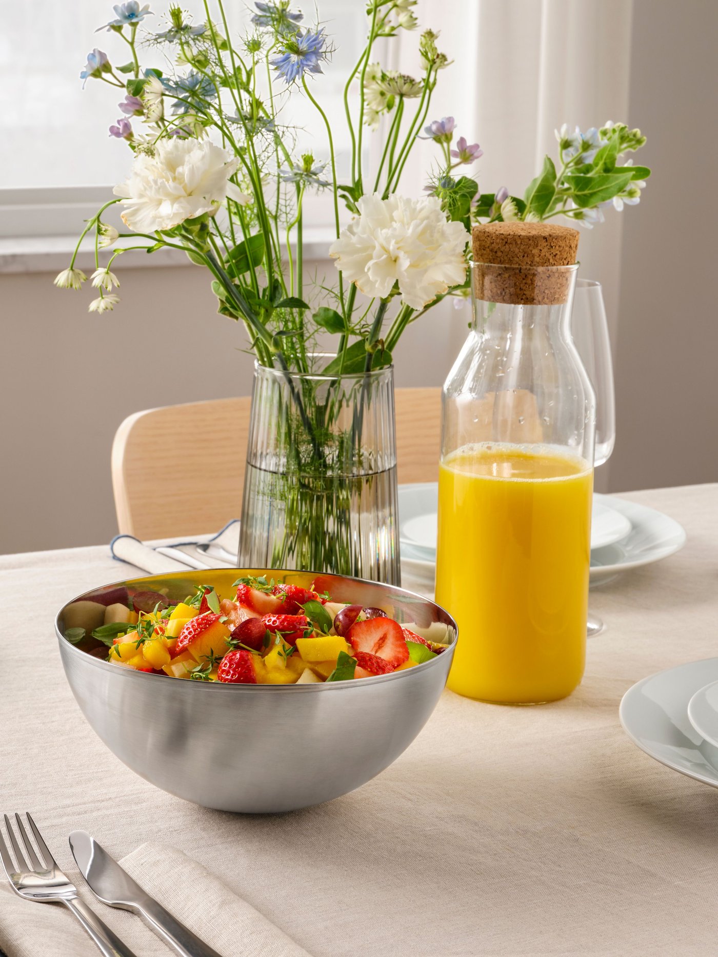 Botella con tapón de corcho IKEA 365+ con jugo de naranja junto a bowl BLANDA con fruta en su interior en una mesa de comedor.