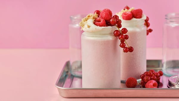 Two light pink LÅNGLUR fruit smoothie milkshakes are topped with red raspberries, red currants and cookie crumbs.