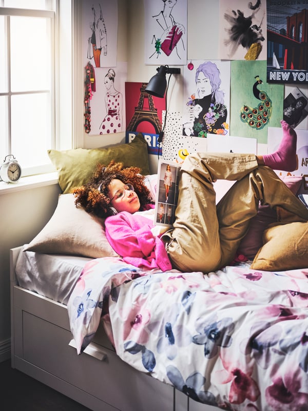 A person lying on a white BRIMNES day bed with two drawers and cushions, and a wall-mounted HEKTAR spotlight above them.